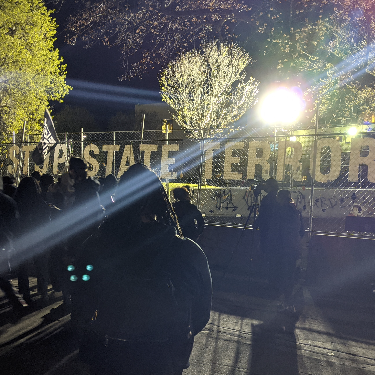 Protesters standing outside police fencing at night. Fencing has letters on it that read Stop State Terror. A bright light from the police shines towa